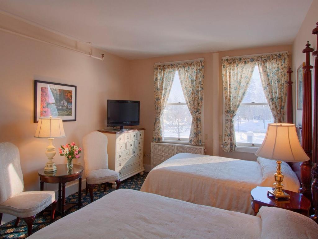 A room with two double beds. Floral curtains hang from the windows and two chairs and a small table sit at the front of the room with a tv to the side of it. 